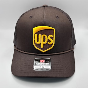 May include: Brown trucker hat with a brown and yellow UPS logo embroidered on the front. The hat has a black mesh back and a black adjustable strap with a red "Richardson" label.