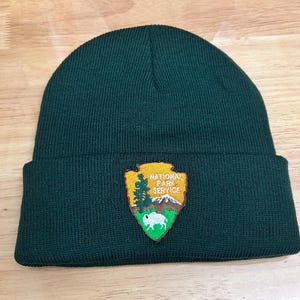 May include: A dark green knit beanie with a brown, green, and white National Park Service patch. The patch features a mountain, a tree, and a bison.