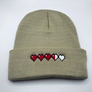 May include: A tan beanie hat with a folded brim. The hat features an embroidered design of four pixelated hearts, three red and one white, representing a health bar.