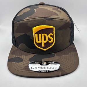 May include: A camouflage baseball cap with a brown UPS logo and a black mesh back. The cap has a black brim and a black and white label that says "SINCE 2000 CAMBRIDGE PB260".