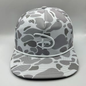 May include: A five-panel baseball cap featuring a white and gray camouflage pattern. The front displays a white embroidered Disney logo. A white cord runs horizontally across the base of the crown, just above the flat brim of the hat.