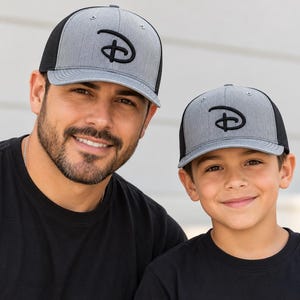May include: Two baseball caps featuring a heather gray front panel and a black mesh back. Each cap displays a prominent black embroidered letter D logo on the front. The caps have a structured crown, a stiff brim, and eyelets on the top panels.