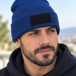 May include: A royal blue knit beanie featuring a rectangular black patch on the front cuff. The beanie is constructed from a ribbed fabric material and has a folded brim design. The patch is solid black and positioned centrally on the front.