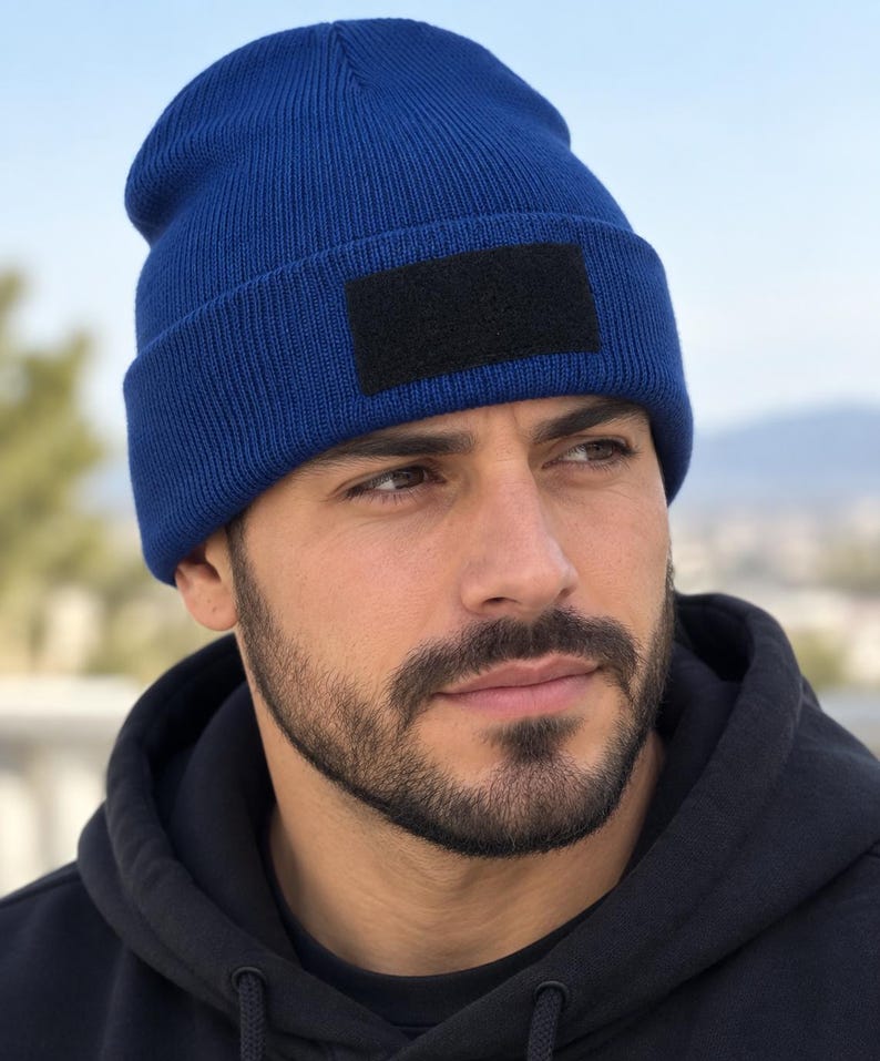 May include: A royal blue knit beanie featuring a rectangular black patch on the front cuff. The beanie is constructed from a ribbed fabric material and has a folded brim design. The patch is solid black and positioned centrally on the front.