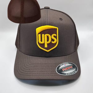 May include: A brown baseball cap with a mesh back and a curved brim. The cap features a yellow and brown UPS logo on the front. The Flexfit label is visible on the brim. The cap is a Flexfit style.