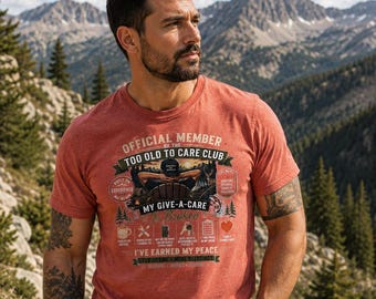 May include: A heathered rust-colored short-sleeve t-shirt featuring a graphic print with the text "OFFICIAL MEMBER OF THE TOO OLD TO CARE CLUB" and "MY GIVE-A-CARE IS BROKEN" along with various small icons including a coffee mug, tools, and a heart.