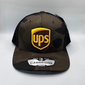 May include: A camouflage-patterned baseball cap with a black mesh back and a curved brim. The front features a yellow and brown UPS logo. The hat has a tag that reads "CAMBRIDGE PB226" and "SINCE 2000".