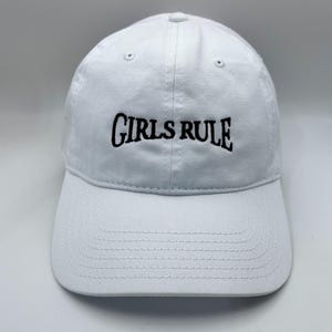 May include: A white baseball cap featuring the text GIRLS RULE embroidered in black thread across the front panel. The cap has a curved brim, a button at the top center, and stitched eyelets for ventilation on the crown panels.