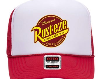May include: A red and white trucker hat featuring a circular logo with the text Medicated Rust-eze Bumper Ointment Rear End Formula in yellow and maroon. The hat has a white foam front panel, red mesh back panels, and a red brim with a small sticker.