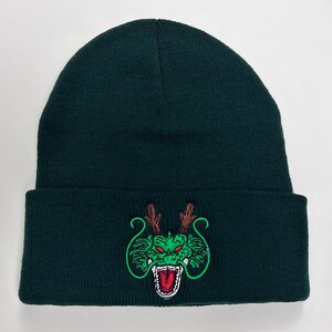 May include: Dark green beanie hat with a folded brim. The hat features an embroidered green dragon design with red accents. The dragon has open jaws and brown antlers. The beanie is made of a knitted material.