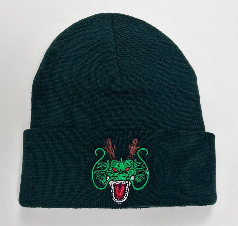 May include: Dark green beanie hat with a folded brim. The hat features an embroidered green dragon design with red accents. The dragon has open jaws and brown antlers. The beanie is made of a knitted material.