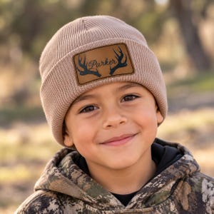 May include: A light brown ribbed knit beanie featuring a rectangular tan leather patch on the front. The patch displays a black graphic of deer antlers flanking the name Parker written in a cursive script font.