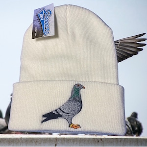 May include: White beanie with a gray embroidered pigeon on the front. The beanie has a fold-over cuff and the brand name "Sluggers Only" is printed on a tag attached to the beanie.