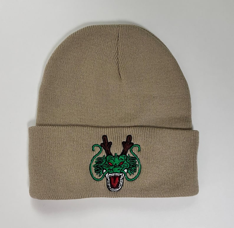 May include: A tan beanie hat with a folded brim. The front of the hat features an embroidered green dragon design with red accents. The hat is made of a soft, knitted material.