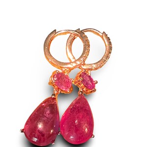 Op de afbeelding: Een paar roségouden oorringen met een kleine ovaal geslepen roze edelsteen aan de bovenkant en een grotere peervormige roze edelsteen als hanger. De ringen zijn aan de buitenrand versierd met kleine heldere steentjes.