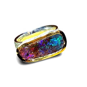 Può includere: Un anello con un inserto ovale allungato in opale iridescente che mostra vivaci riflessi blu, verdi e viola. La pietra è incastonata in una lunetta color oro lucido, in contrasto con una fascia color argento dalla superficie testurizzata.