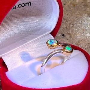Può includere: Un anello aperto con fascia in argento e due castoni dorati, ciascuno contenente una pietra rotonda iridescente con riflessi verdi e blu. L'anello è esposto all'interno di un portagioie in velluto rosso a forma di cuore con interno bianco.