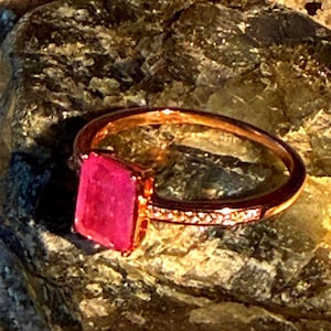Può includere: Un anello in oro rosa con una pietra preziosa rettangolare di colore rosa vibrante incastonata a griffe. La fascia è sottile e metallica, con una finitura lucida che riflette la luce, mettendo in risalto la pietra sfaccettata al centro.