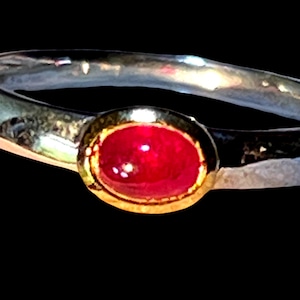 May include: A silver ring featuring a hammered, textured band and a central oval-shaped, cabochon-cut red gemstone set within a gold-toned bezel. The ring has a polished finish on the metal surfaces, highlighting the contrast between the silver band and the gold setting.