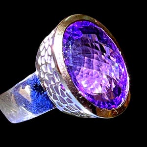 Könnte beinhalten: Ein Silberring mit einem großen, facettierten, oval geschliffenen violetten Amethyst, der in einer Fassung sitzt. Die Ringschiene weist eine strukturierte, schuppenartige Oberfläche auf, die einen Kontrast zum glatten, polierten Metall bildet.