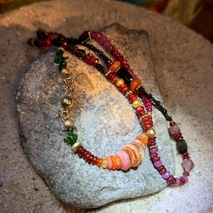 Può includere: Una collana di perline a più fili con un mix vivace di perline in pietra rossa, rosa, arancione, nera e verde. Il design incorpora piccoli distanziatori in metallo dorato e una chiusura magnetica dorata, creando un aspetto a strati con forme, dimensioni e texture delle perline variegate lungo l'intera lunghezza del pezzo.