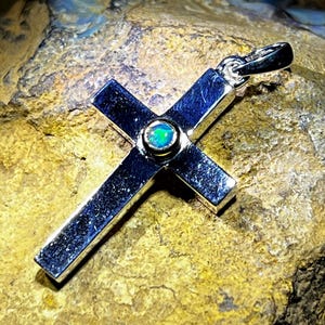 Könnte beinhalten: Ein silberner Kreuzanhänger mit einem kleinen, runden, schillernden Opalstein in der Mitte. Das Kreuz hat eine polierte, reflektierende Oberfläche und eine Schlaufe für eine Kette. Der Anhänger wird auf einer strukturierten, goldbraunen Oberfläche präsentiert.