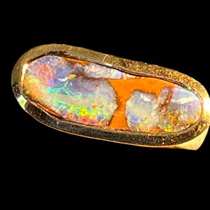 Könnte beinhalten: Ein länglicher, ovaler Ring mit einer Boulder-Opal-Einlage, die lebhafte blaue, grüne und rote Feuerblitze aufweist und in ein dunkles, strukturiertes Metallband gefasst ist. Der Stein zeigt natürliche, erdige braune Matrix-Abschnitte.