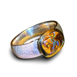 Op de afbeelding: Een ring met een textuur zilveren band en een in een zetting geplaatste ovale steen. De steen heeft een warme bruine basiskleur met levendige, iriserende flitsen van blauw, groen en oranje, omlijst door een gepolijste goudkleurige zetting.