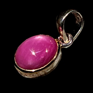 May include: A silver pendant featuring a large, oval magenta gemstone. The gemstone is set in a silver bezel with a gold-coloured rim. The pendant has a loop at the top for a chain.