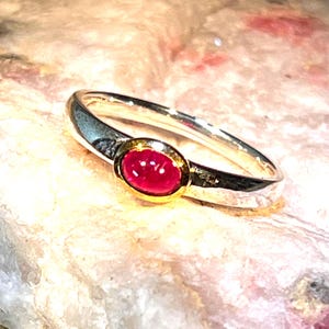 Könnte beinhalten: Ein Silberring mit einem glatten, ovalen, tiefrosa Cabochon-Edelstein, der in einer goldfarbenen Fassung sitzt. Das Ringband besteht aus poliertem Silber und der Stein ist horizontal zentriert, was einen Kontrast zwischen Silber und Gold erzeugt.