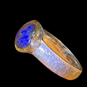 Könnte beinhalten: Ein Ring mit einem kreisförmigen, tiefblauen Lapislazuli-Stein in einer goldfarbenen Fassung. Die Ringschiene hat eine texturierte, matte Oberfläche mit metallischem Glanz, die einen Kontrast zur glatten, polierten Oberfläche des Edelsteins bildet.