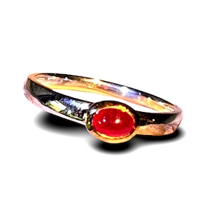 Op de afbeelding: Een gouden ring met een enkele, kleine, ovaalvormige rode edelsteen in een gladde zetting. De band heeft een gepolijste, reflecterende metalen afwerking die het licht opvangt, waardoor heldere accenten op het gebogen oppervlak ontstaan.
