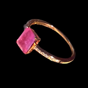 Könnte beinhalten: Ein goldener Ring mit einem quadratisch geschliffenen, leuchtend rosa Edelstein in der Mitte. Die Ringschiene ist schmal und mit einer Reihe kleiner, klarer, funkelnder Steine besetzt, die an der Seite eingelassen sind und für ein elegantes Design sorgen.