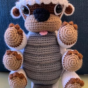 May include: A crocheted stuffed animal resembling Appa from Avatar: The Last Airbender. The animal is white with brown accents and has a gray body. It has large, round eyes and a pink tongue sticking out.