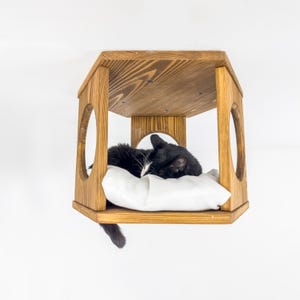 May include: A black and white cat sleeps on a white cushion inside a wooden cat bed. The bed is hexagonal in shape and has a wooden frame with rounded cutouts. The bed is suspended from the ceiling.