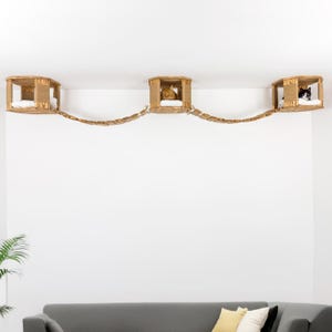 Puede incluir: Un conjunto de muebles para gatos con tres casas de madera conectadas por un puente de cuerda. Cada casa tiene un cojín blanco. Se ven dos gatos descansando en las casas. Las casas para gatos están montadas en una pared blanca.