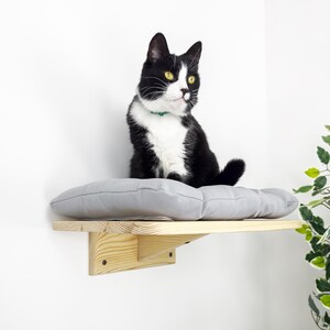 May include: A black and white cat sits on a gray cushioned shelf attached to a wall. The shelf is made of light wood and has a simple design.