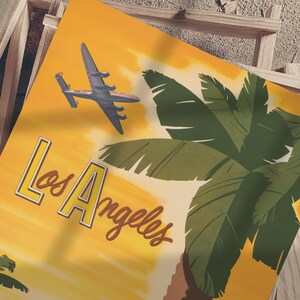 Puede incluir: Un cartel de viaje vintage con fondo amarillo que presenta una palmera y un avi&oacute;n volando sobre una puesta de sol. El texto "Los Angeles" est&aacute; escrito en una fuente retro.