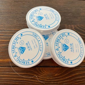 以下が含まれることがあります： 木製の表面に重ねられた、ジュエリー作りに使用する白いストレッチ糸の複数のスプール。各スプールには、日本語のテキスト、ダイヤモンドのグラフィック、および「STRONG & STRETCHY」という単語が記載された青と白のラベルが付いています。
