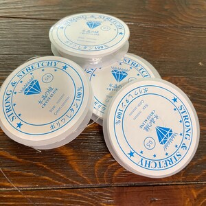 以下が含まれることがあります： 透明で伸縮性のあるビーズコードの複数のスプール。各スプールには、青い文字とダイヤモンドのグラフィック、日本語のテキストが入った白いラベルが付いています。ラベルには「Strong & Stretchy」と記載されています。コードの太さは0.5mmです。