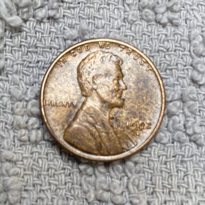 May include: A copper-colored 1992 United States penny. The obverse features a profile of Abraham Lincoln, with the words "LIBERTY" and "IN GOD WE TRUST" visible. The year "1992" is also displayed.