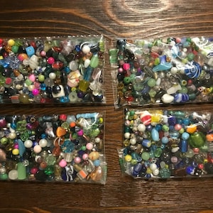 May include: Four clear plastic bags filled with a variety of colorful beads. The beads are in various shapes and sizes, including round, square, and oblong. Colors include red, green, blue, yellow, and clear. The bags are arranged on a dark brown wooden surface.