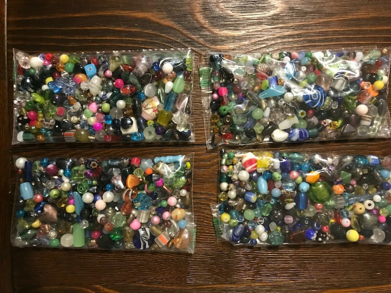 May include: Four clear plastic bags filled with a variety of colorful beads. The beads are in various shapes and sizes, including round, square, and oblong. Colors include red, green, blue, yellow, and clear. The bags are arranged on a dark brown wooden surface.