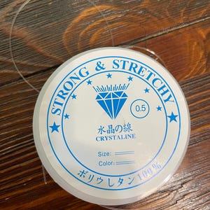 以下が含まれることがあります： 透明で伸縮性のあるビーズコードのスプール。白いラベルには「STRONG & STRETCHY」と「CRYSTALINE」という青い文字が書かれています。ラベルには日本語のテキストと数字「0.5」も含まれています。