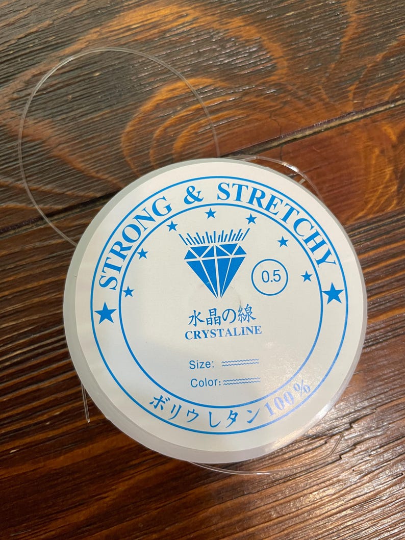 以下が含まれることがあります： 透明で伸縮性のあるビーズコードのスプール。白いラベルには「STRONG & STRETCHY」と「CRYSTALINE」という青い文字が書かれています。ラベルには日本語のテキストと数字「0.5」も含まれています。