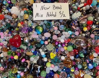 May include: A vibrant assortment of beads in various shapes, sizes, and colors, including red hearts, clear crystals, and iridescent pieces. A handwritten sign reads "New Bead Mix Added 5/2". The beads are scattered, creating a colorful, textured display.