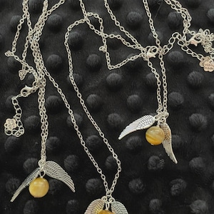 May include: Three silver-toned necklaces with a chain design. Each necklace features a pair of detailed wings and a round, yellow-brown gemstone. The necklaces are displayed on a black, textured surface.