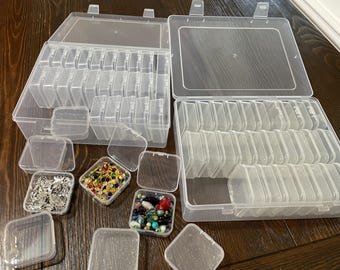 May include: Clear plastic storage containers with multiple compartments, ideal for organizing small items. Several open containers are visible, some filled with beads and jewelry findings. The containers are on a wooden surface.