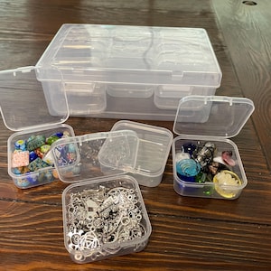 May include: Clear plastic organizer with multiple compartments and several smaller, individual containers. The smaller containers hold various craft supplies, including colorful beads and silver charms. The organizer is on a wooden surface.