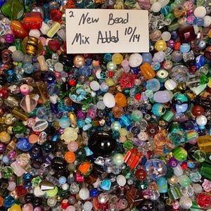 May include: A large collection of assorted beads in various shapes, sizes, and colors, including clear, opaque, and metallic finishes. A white rectangular card is placed on top with the handwritten text 2 New Bead Mix Added 10/14.
